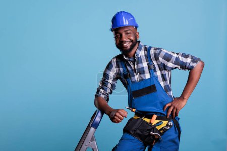 Photo for Builder looking happy and relaxed at work, dressed in coveralls and protective helmet leaning on portable ladder. Optimistic construction worker wearing tool belt while taking a break. - Royalty Free Image