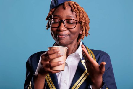 Téléchargez les photos : Professional female flight attendant delighted with scent of freshly brewed coffee during break time at work. Air hostess dressed in work uniform against blue background. - en image libre de droit