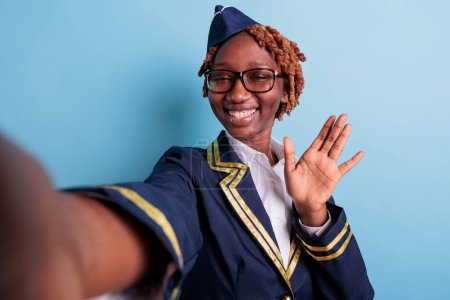 Photo for African american flight attendant waving happily in studio selfie. Aviation worker wearing glasses, afro woman taking self portrait with cellphone, smartphone. Stewardess portrait greeting. - Royalty Free Image