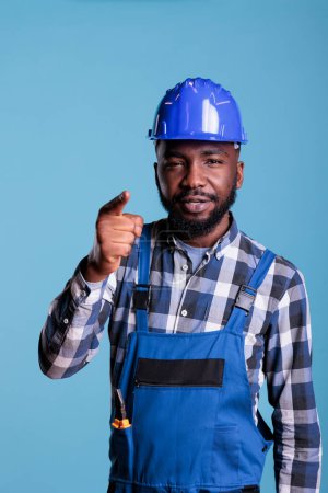 Photo for African american construction worker pointing index finger at camera and explaining thoughts. Builder with authority having conversation talking about decisions, studio shot against blue background. - Royalty Free Image