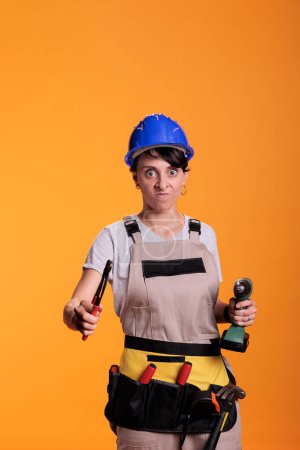 Foto de Mad crazy woman contractor posing with renovating tools, holding pair of pliers and drilling nail gun in studio. Angry construction worker acting stressed with drill over yellow background. - Imagen libre de derechos