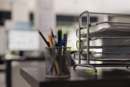 Foto de Close up view of metal pencil holder with colored pencils in creative design office, next to folder tray with project documents. Out of focus shot of monitor on in background in empty workplace. - Imagen libre de derechos