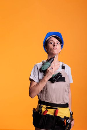 Foto de Female repair contractor holding power drill in front of camera, using electric drilling tool before renovation project. Construction worker wearing blue helmet and building uniform. - Imagen libre de derechos