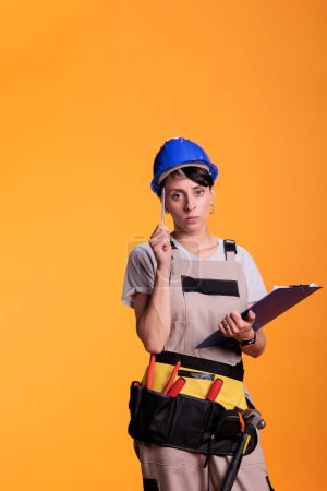 Photo for Female builder brainstorming ideas with clipboard papers in studio, taking notes on files and thinking about renovating solution. Industrial engineer using measurements to build site. - Royalty Free Image