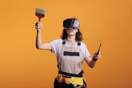 Photo for Woman construction worker wearing vr glasses while she holds paint brush in studio. Female contractor with overalls using virtual reality headset before painting walls for renovation project. - Royalty Free Image