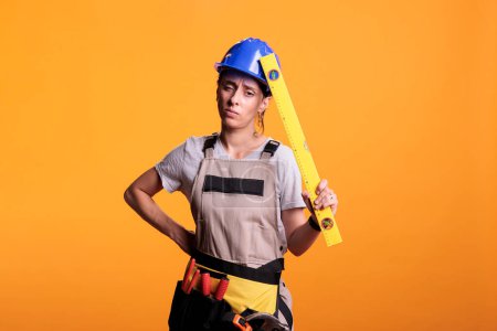 Photo for Construction worker suffering from painful headache and holding leveler on camera, rubbing temples. Woman renovation expert with migraine dressed with overalls and hardhat in studio. - Royalty Free Image