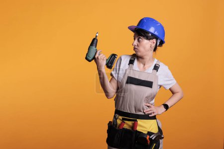 Foto de Disappointed woman contractor holding electric nail gun, posing over background. Female builder wearing overalls and hardhat in studio, using poer drilling tool for maintenance. - Imagen libre de derechos