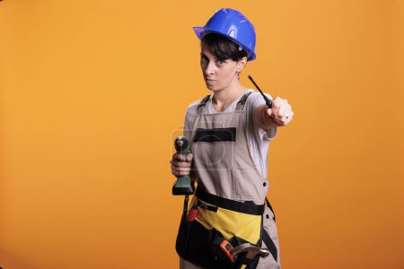 Photo for Focused female contractor holding drilling gun and screwdriver, posing in studio. Woman dressed as building worker holding renovating tools and power screw drill gun, serious builder. - Royalty Free Image