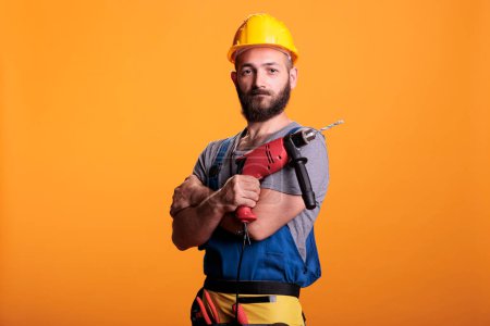 Photo for Happy building contractor holding power drill gun and posing in studio, looking at camera. Craftsman builder using drilling nail tool wearing uniform and hardhat, renovating service. - Royalty Free Image