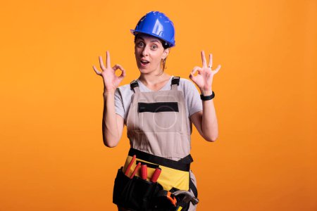 Téléchargez les photos : Young female contractor doing okay good symbol in studio, wearing renovating uniform and helmet. Cheerful construction worker showing approval and agreement gesture, achievement. - en image libre de droit