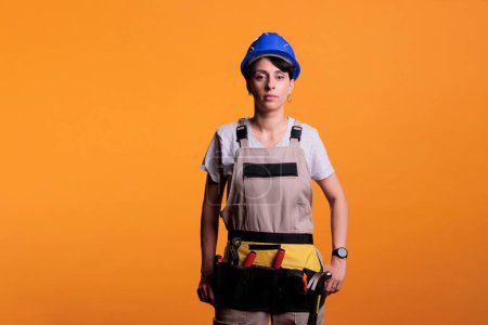 Téléchargez les photos : Young contractor wearing protective hardhat and looking confident at camera in studio. Confident woman construction worker posing, working in refurbishment industry over yellow background. - en image libre de droit
