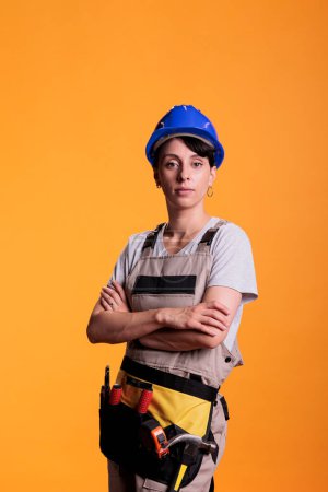Photo for Portrait of professional builder posing with arms crossed in studio. Professional woman in construction industry looking confident, standing in renovation overalls over yellow background. - Royalty Free Image