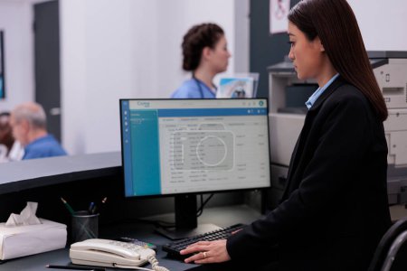 Foto de Receptionist standing at hospital counter checking medical expertise on computer while planning checkup visit appointments for patients. Medicine support service and concept - Imagen libre de derechos