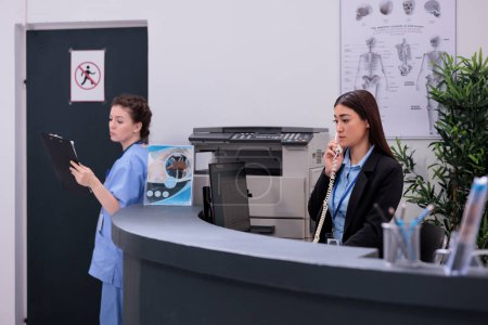 Photo for Asian receptionist standing at reception counter talking at landline phone with remote patient discussing medical expertise. Assistant holding clipboard analyzing disease report in waiting area - Royalty Free Image