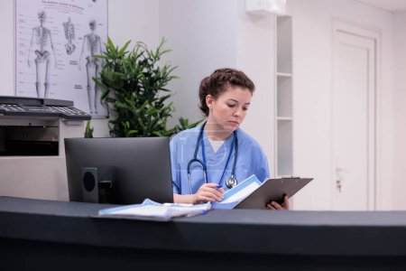 Foto de Caucasian nurse working at reception counter looking at medical expertise while planning patients checkup visits. Health care professionals are trained to diagnose and treat various illnesses. - Imagen libre de derechos