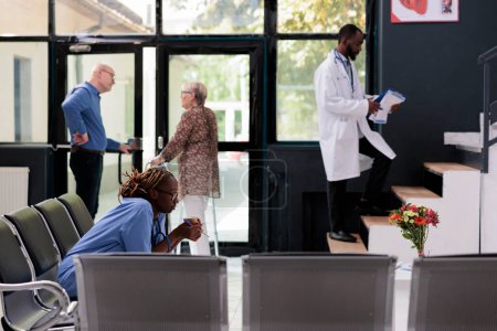 Téléchargez les photos : African american nurse sitting on chair in hospital lobby being tired during consultation, having a break before start working at patient health care treatment. Medicine service and concept - en image libre de droit