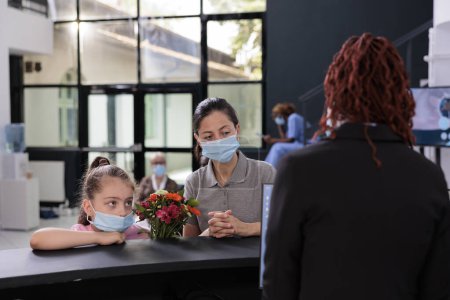 Photo for Mother and child wearing protective face mask against covid19 while discussing medical insurance with receptionist during checkup visit examination in hospital lobby. Medicine service and concept - Royalty Free Image