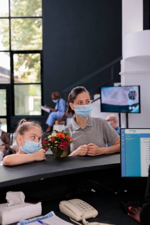 Foto de Receptionist talking with mother and child explaining medical expertise during checkup visit appointment in hospital reception. People wearing protective face mask to prevent infection with covid19 - Imagen libre de derechos