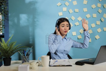 Foto de Company employee wearing wireless headset attending a virtual business meeting raising her hand to give an opinion. Professional woman sitting at desk in workplace on video call with colleagues. - Imagen libre de derechos