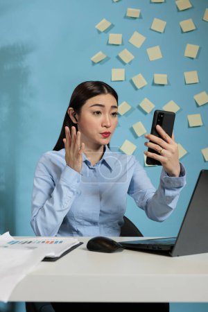 Photo for Woman having a video conference with coworkers while sitting in a modern office. Professional female worker wearing a wireless earphone and speaking in an online meeting. - Royalty Free Image