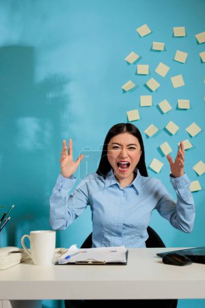 Foto de Female corporate employee screaming and waving her arms sitting at modern office desk with laptop. Woman feeling exhausted by demands of boss in new project she is working on. - Imagen libre de derechos