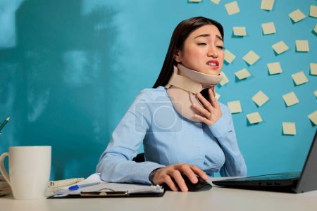 Téléchargez les photos : Asian businesswoman with medical foam cervical collar after physical injury and accident. Corporate employee working with pain and discomfort sitting at modern office desk. - en image libre de droit