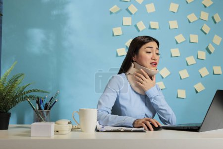 Photo for Asian woman wearing cervical collar to recover from injury. Office employee with fractured vertebrae wearing medical neck brace while working from desk to finish marketing reports. - Royalty Free Image