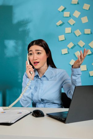 Foto de Corporate female employee sitting at a desk in the workplace holding a mobile phone makes a business call and listens to customer complaints feeling upset. Woman gesturing with hands in office. - Imagen libre de derechos