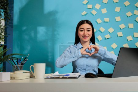 Photo for Friendly professional woman making a heart shaped sign indicating positivity and love. Corporate female employee sitting at office desk showing positive feelings while working. - Royalty Free Image