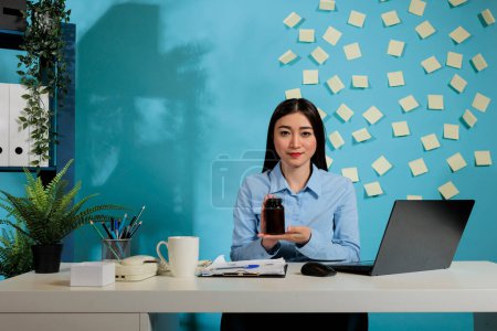 Foto de Corporate female employee at contemporary office desk with dark container of pills looking at camera. Professional woman showing capsules bottle with blue wall in background. - Imagen libre de derechos