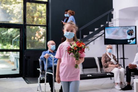 Téléchargez les photos : Kid standing in hospital waiting area while holding bouqet of flowers during checkup visit consultation. Multi ethnic people wearing protective medical face mask to prevent infection with coronavirus - en image libre de droit