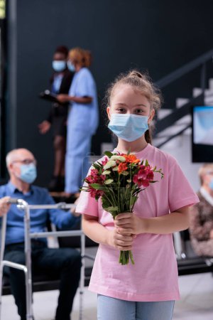 Téléchargez les photos : Little girl looking at camera while holding bouquet of flowers waiting for grandmother to finish consultation. People wearing medical face mask to prevent infection with covid19 in hospital area - en image libre de droit