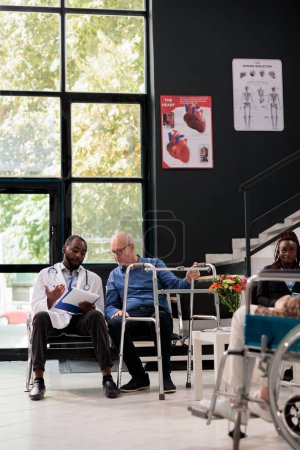 Foto de African american doctor holding clipboard explaining medical expertise to senior patient with wallking frame during consultation in hospital waiting area. Medicine service and concept - Imagen libre de derechos