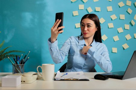 Foto de Professional woman sitting at office desk on video call with business partner looking at mobile phone screen. Corporate employee at startup workstation using electronic device. - Imagen libre de derechos