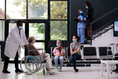 Téléchargez les photos : Physician doctor bringing grandmother in wheelchair to family after medical consultation during checkup visit appointment. Granddaughter holding bouqet flowers for senior woman. Virus epidemic - en image libre de droit