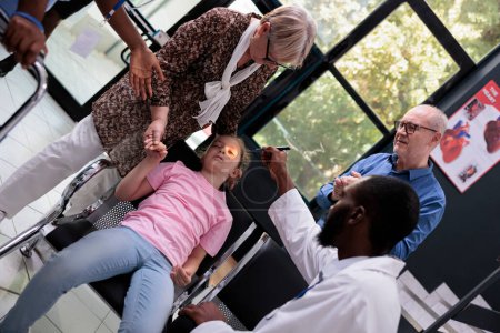 Téléchargez les photos : African american medic helping girl child after fainting on chair in hospital reception during checkup visit consultation. Grandparents being worried trying to help granddaughter. Medicine service - en image libre de droit