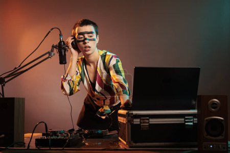 Téléchargez les photos : Happy musician playing with techno music on mixer, having fun with turntables as disc jockey and mixing sounds in nightclub studio. Using headphones and microphone to test bass control key. - en image libre de droit