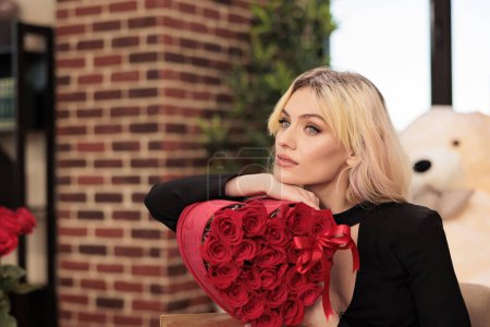 Foto de Beautiful woman posing lovely with red roses bouquet, celebrating valentines day at home in living room. Attractive blonde girlfriend receving flowers in heart shaped box, love holiday event. - Imagen libre de derechos
