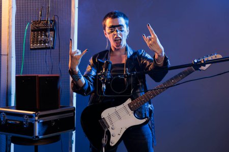Foto de Crazy musician doing rock sing with fingers and screaming loud in studio, playing guitar and singing heavy metal music. Cool alternative artist performing punk rock song, wearing leather. - Imagen libre de derechos