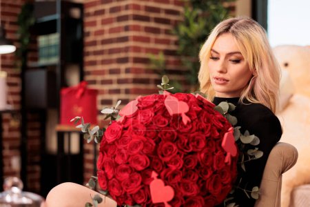 Téléchargez les photos : Beautiful woman holding flower bouquet in heart shaped box, enjoying celebrating valentine s day. Attractive blonde girlfriend with in black dress standing in living room filled with romantic presents - en image libre de droit