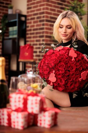 Photo for Cute girl standing in living room filled with valentines day gifts, holding red roses large bouquet. Beautiful blonde girlfriend in elegant black dress enjoying celebrating love holiday - Royalty Free Image