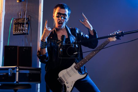 Téléchargez les photos : Punk rocker doing rock sign on live performance, singing loud alternative heavy metal music. Using microphone and guitar to play cool solo song, wearing leather jacket and showing rock on symbol. - en image libre de droit