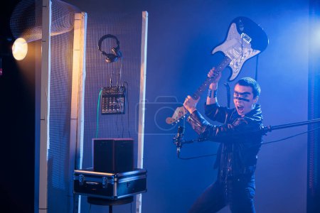 Foto de Ecstatic artist holding bass guitar to smash and act crazy, performing punk rock music and screaming loud in studio. Rocker with leather jacket fooling around and throwing musical instrument. - Imagen libre de derechos