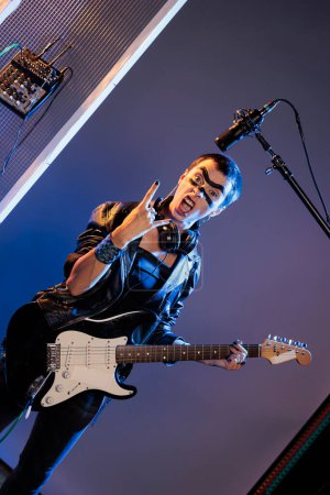 Téléchargez les photos : Punk rocker doing rock sign and playing guitar, feeling ecstatic and doing solo concert in studio over background. Female singer showing rock and roll symbol at live show performance. - en image libre de droit