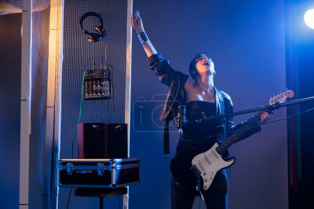 Téléchargez les photos : Punk rocker doing live performance in studio, creating funky cool vibe with heavy metal alternative music. Stylish female artist singing loud songs and acting ecstatic in studio background. - en image libre de droit