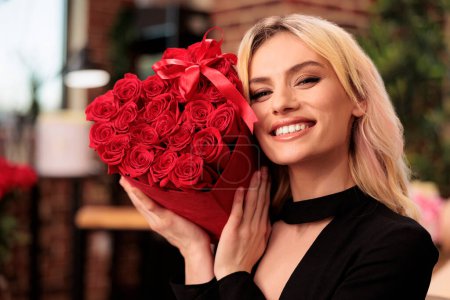 Téléchargez les photos : Girlfriend holding red roses bouquet celebrating love holiday, standing in living room filled with valentine s day presents. Attractive cute woman in elegant black dress enjoying romantic gift. - en image libre de droit