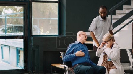Téléchargez les photos : African american people helping elderly man with heart problems to sit in hospital reception lobby. Senior physician consulting old patient with stethoscope, trying to cure cardiovascular illness. - en image libre de droit