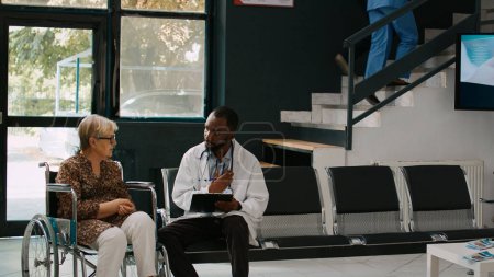 Téléchargez les photos : Médecin afro-américain consultant une vieille femme en fauteuil roulant, parlant de handicap physique et de maladie dans la salle d'attente de l'hôpital. Spécialiste et patient ayant une déficience faisant un examen médical. - en image libre de droit