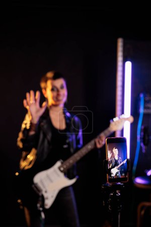 Téléchargez les photos : Smiling rockstar greeting fans while playing at electric guitar in front of phone camera in sound studio, working at rock album using electricinstrument. Rebel musician recording heavy metal song - en image libre de droit