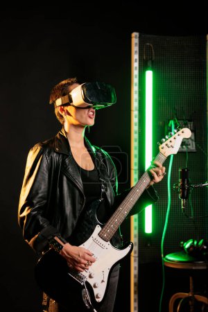 Photo for Woman performer playing at electric guitar in sound studio while wearing vr goggles for concert simulation, enjoying music performance. Rebel performer working at new heavy metal album - Royalty Free Image